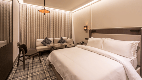 Modern hotel room at The Residency Towers Chennai featuring a large bed, seating area with sofa and chair, patterned carpet, and warm ambient lighting.
