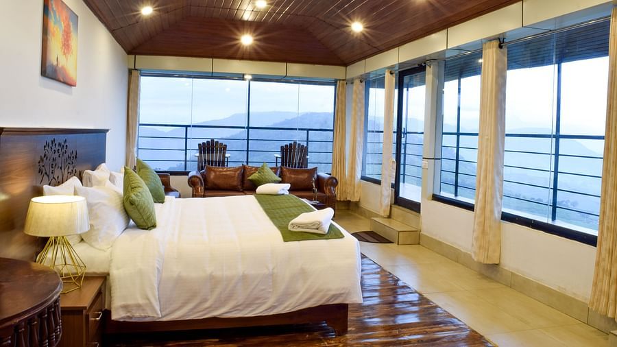 A side angle shot of the suite at the resort feature a expansive balcony that overlooks the lush green valleys, king size bed with white bed sheets and a green bed runner  | Ibex Stays and Trails