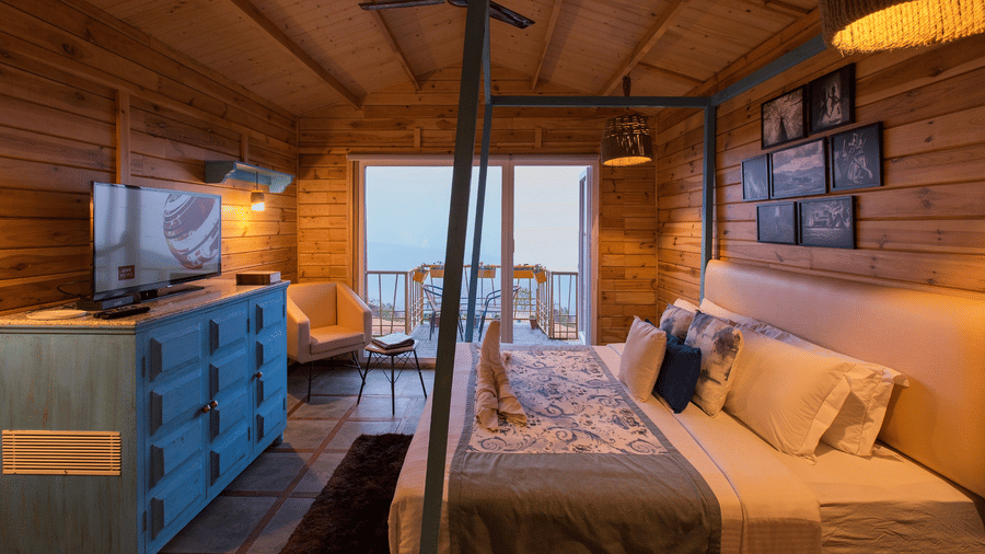 A bedroom at Polo Cherrapunjee Resort with a bed, a colourful dresser, seating area, and glass doors opening to a balcony with scenic views.