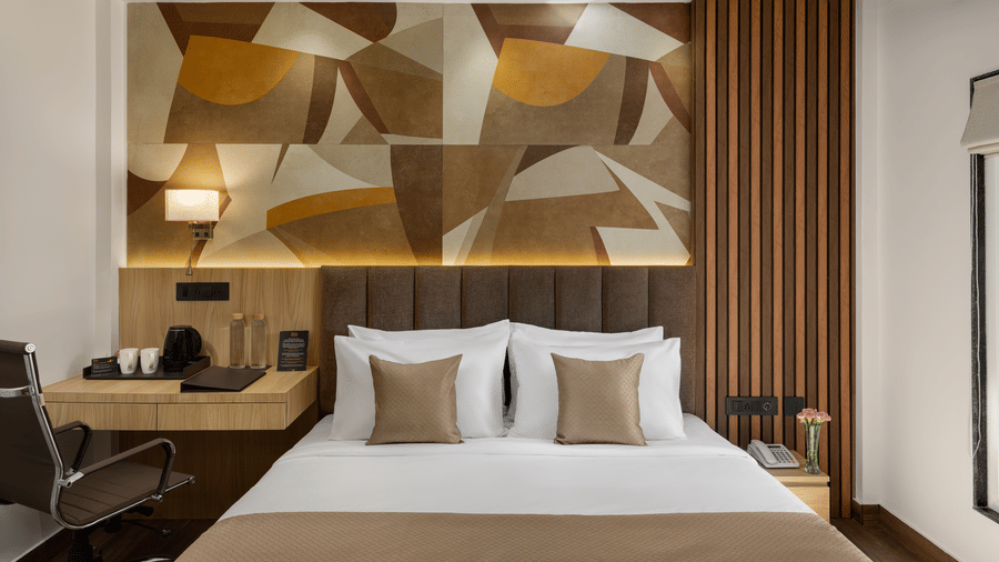 A brightly lit hotel room showing a large double bed with light bedding, a desk and chair on the left, and a headboard wall featuring a geometric pattern | Voyage Eastin Suites