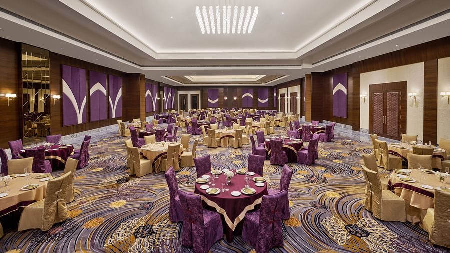 A spacious, elegant banquet hall with round gold-draped tables, purple and gold chairs, and refined place settings. The high ceiling features recessed lighting, a modern chandelier, and purple-gold accents with wood panelling - Voyage The Solitaire Bliss.