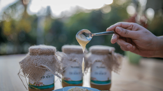 A close up shot of 3 sealed jars of honey with a bowl of honey being poured by a person - Karma Lakelands.