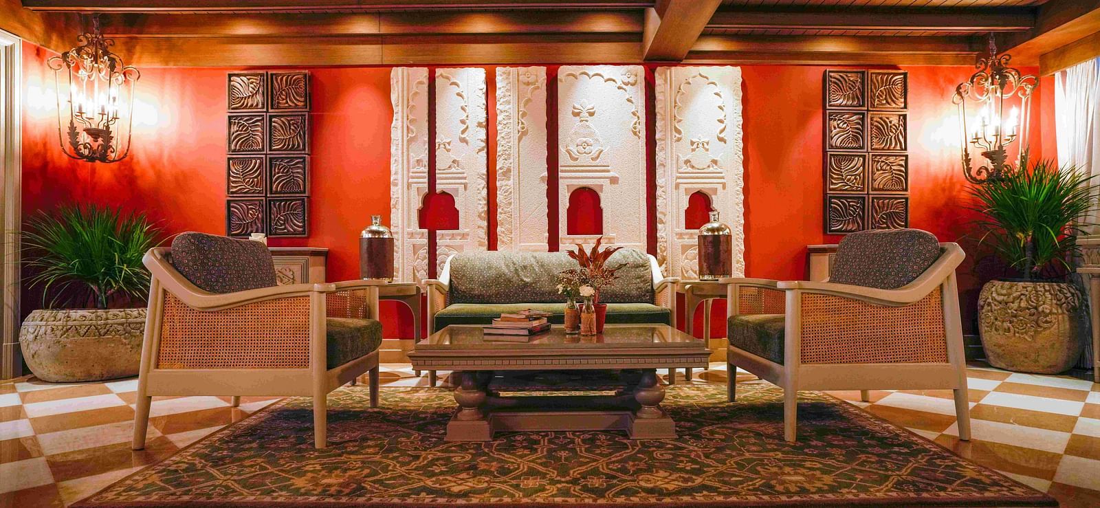 A wide shot of a welcoming lounge area at The Evren, Vagator. The space features a green sofa and two chairs with cane details, set against a vibrant red wall with ornate white carvings. The room has a checkered floor and a decorative rug.