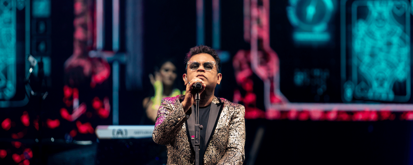 A. R. Rahman performing live on stage, singing into the microphone with vibrant lights and digital graphics in the background.