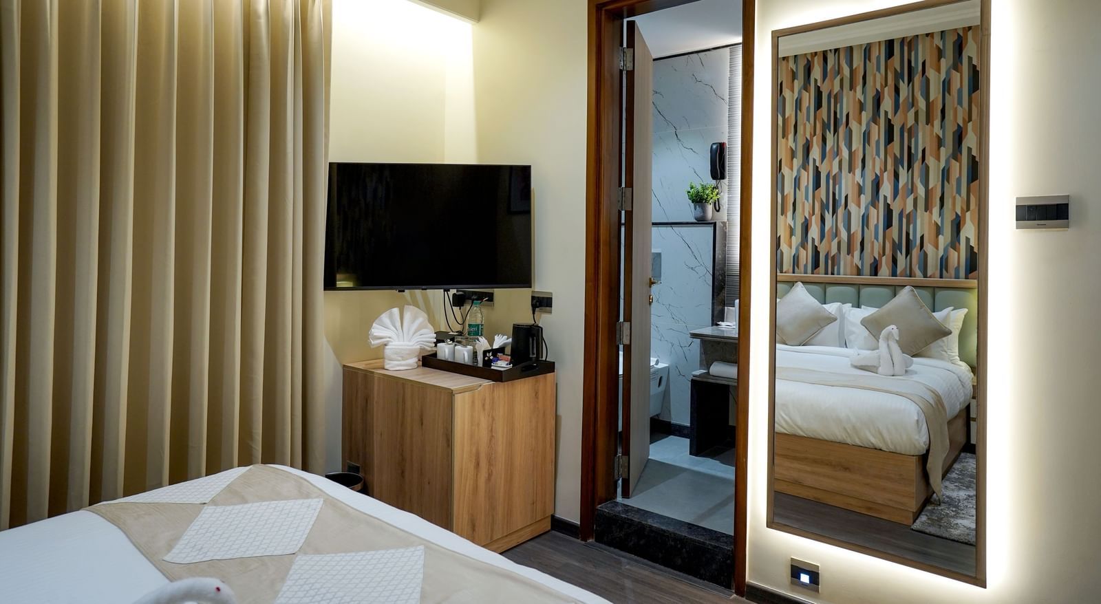 A wall-mounted television alongside a door leading to the bathroom and a full length mirror in the Deluxe Room at M2 Square by Monday Premium.