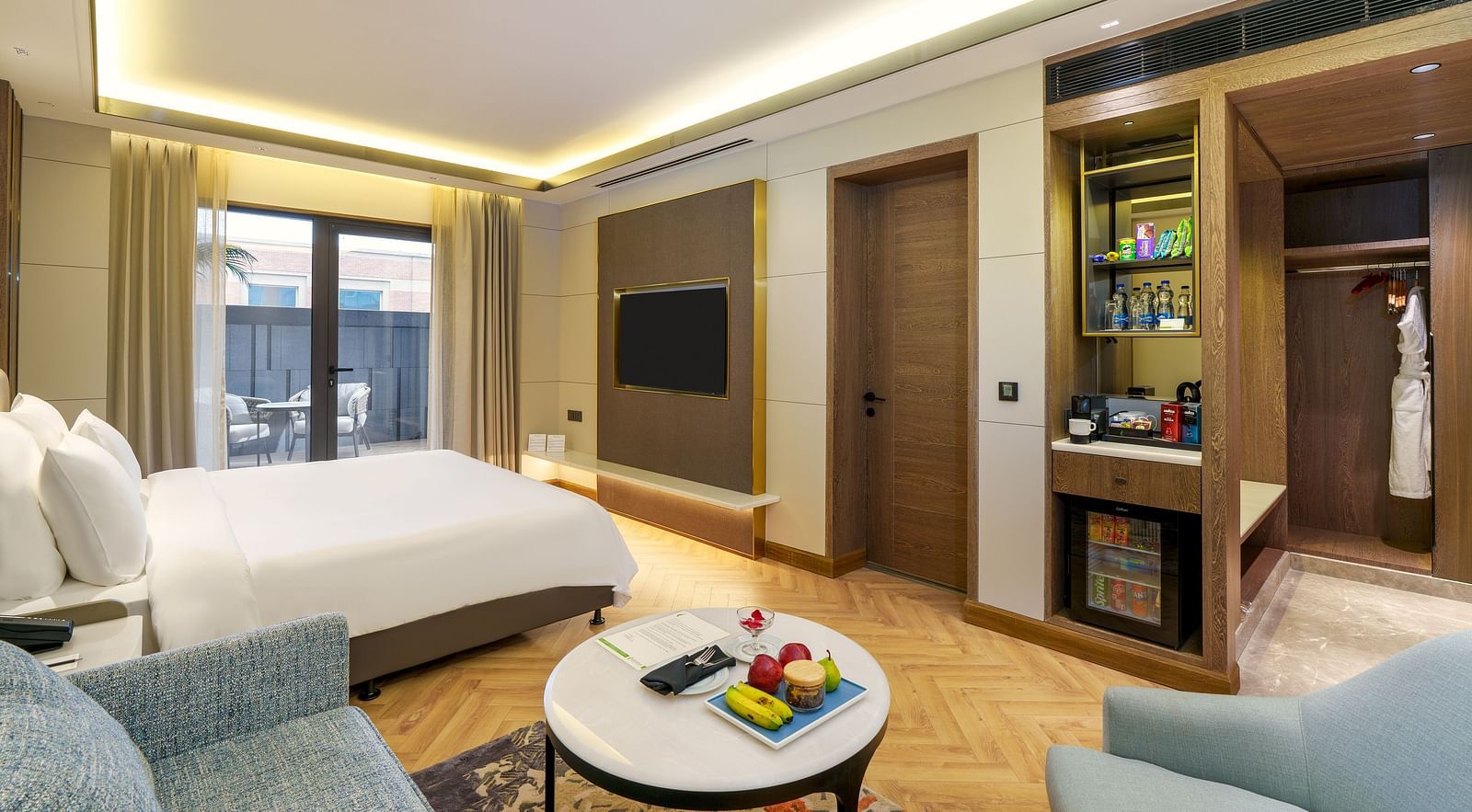 A modern hotel suite at Nirvana Luxury Hotel, Ludhiana, showcasing a bed, a plush blue sofa, a round table with amenities, a wall-mounted TV, and access to a balcony.