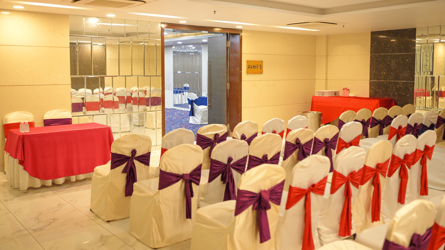 A well lit function venue at Ambar Sarovar Portico, Gandhidham includes chairs with decorative bows, a side table, and a wide open doorway.