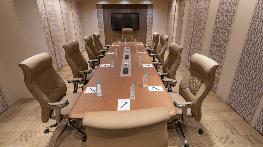 A brightly lit boardroom featuring a long wooden table with multiple swivel chairs, a wall-mounted television screen, and patterned walls | Pride Elite Chhatrapati Sambhajinagar