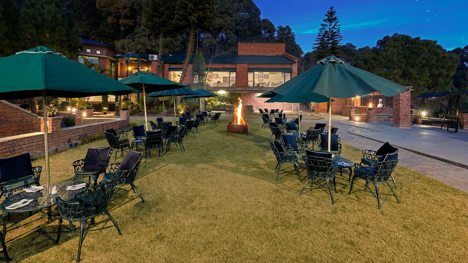 An outdoor lawn with an umbrella-shaped roofs providing shade, surrounded by chairs arranged for seating -  Baikunth resort, kasauli 