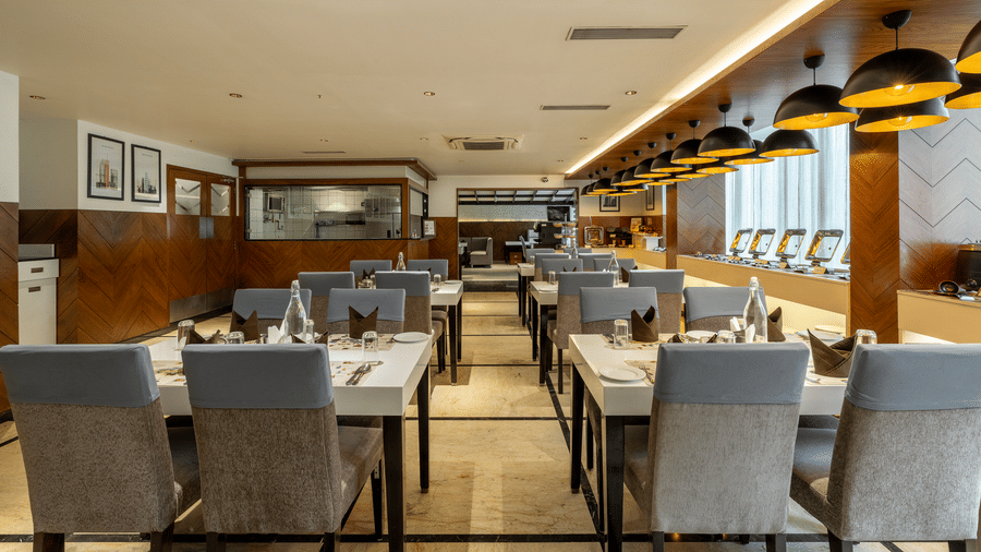 Restaurant dining area at The Citrine, Bangalore, with set tables, grey upholstered chairs, framed art on wood-panelled walls, and chafing dishes behind.