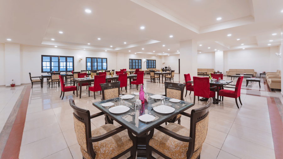 An indoor dining area Shri Vallabh Vilas Lords Plaza Nathdwara featuring red and beige coloured chairs with wooden furnishings, clean tile floor and rows of windows.