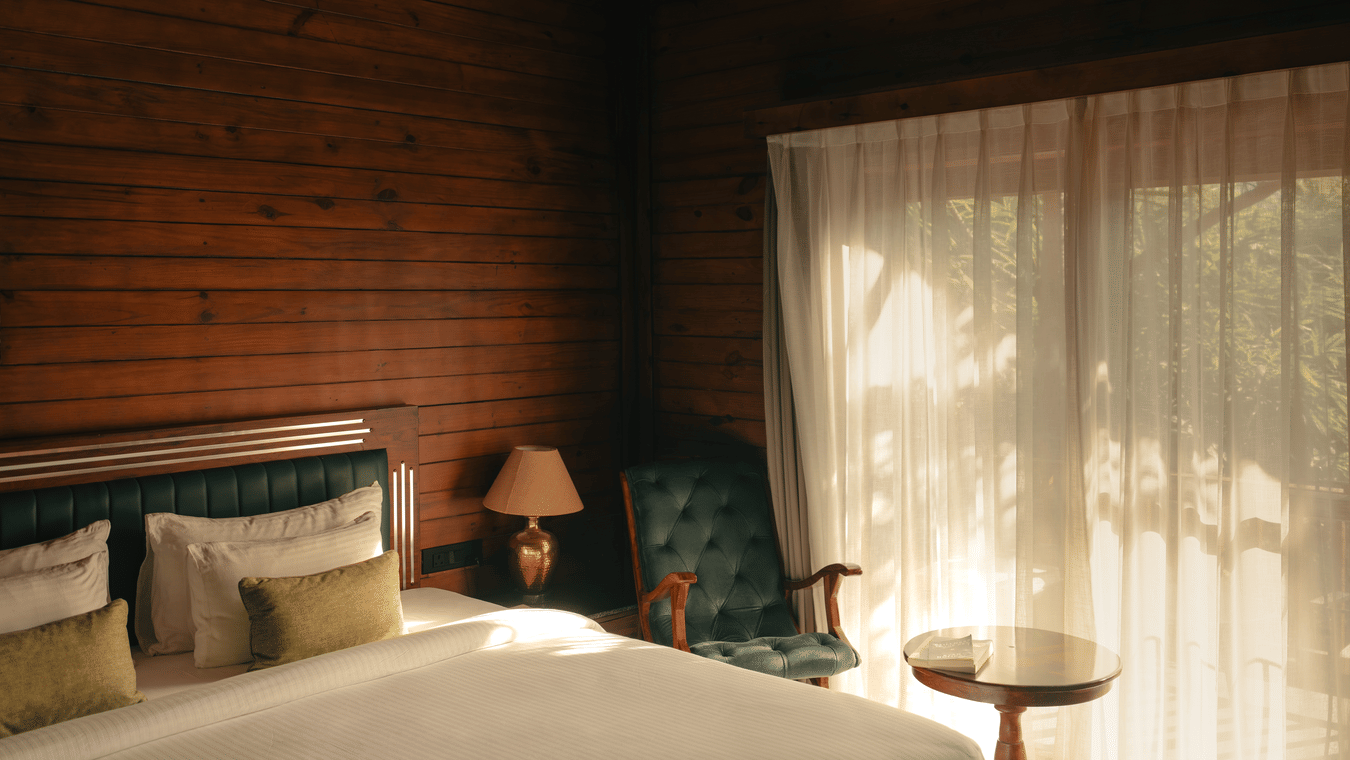 A bedroom at The Ananta Udaipur featuring a king size bed, side table, armchair, small round coffee table, and large window with curtains.