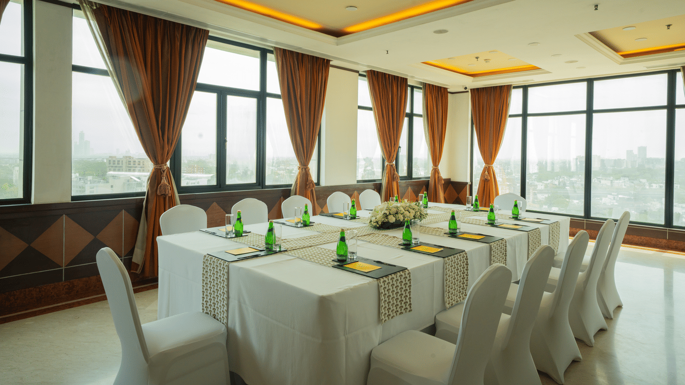 Sampan Tea Room at The Suryaa set with rectangular tables, white covers, green bottles, notepads, pens, warm recessed lighting, large windows with city view and brown curtains.