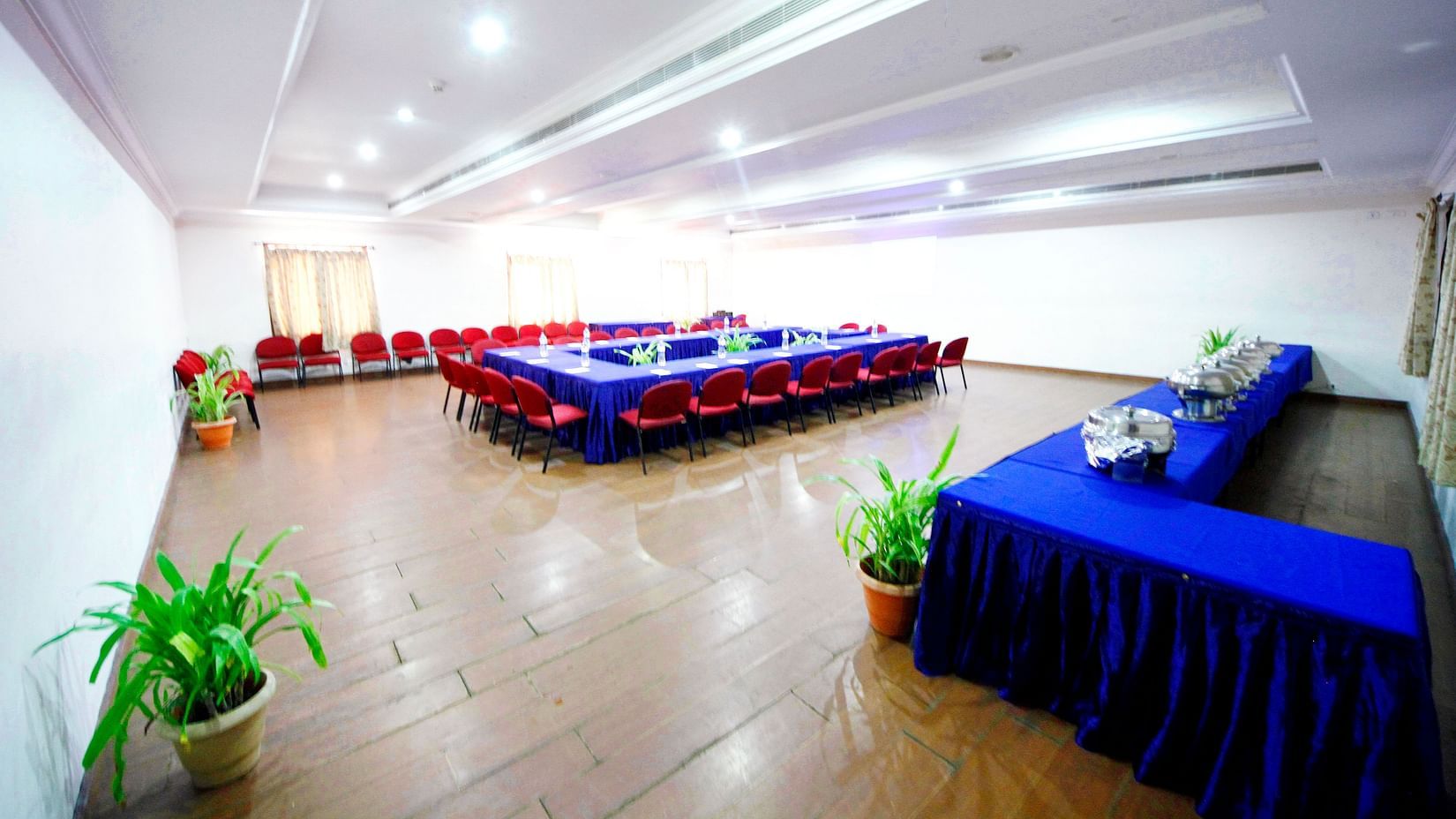 A spacious Pearl hall featuring a U-shaped table arrangement, buffet setup to the right, wooden floor, potted plants, and with windows and curtains
