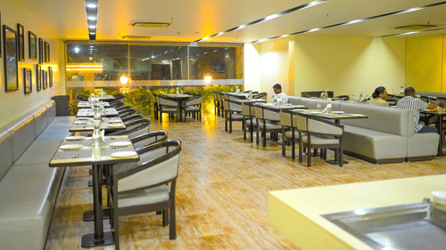 A restaurant dining room at Ambar Sarovar Portico, Gandhidham features neatly arranged tables, chairs, and cushioned booth seating along the wall.