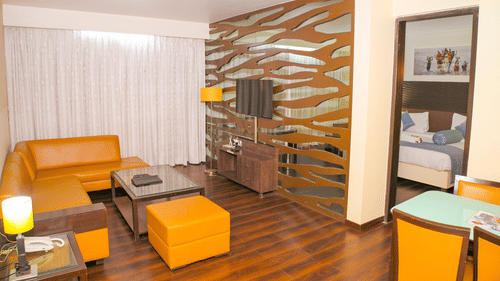 A living room area of a suite at Ambar Sarovar Portico, Gandhidham features a wooden floor, a television, sofa seating, and a dining table.