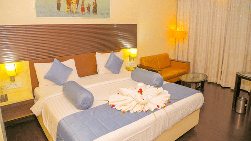 A hotel bedroom at Ambar Sarovar Portico, Gandhidham shows a large king size bed decorated with folded towels, a sofa, and a wall painting.
