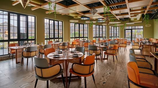 Vibrant and spacious dining area with floor-to-ceiling windows, natural light flooding in, earthy tones with orange and grey seating, wooden lattice ceiling with hanging planters and geometric lights - Voyage The Solitaire Bliss.