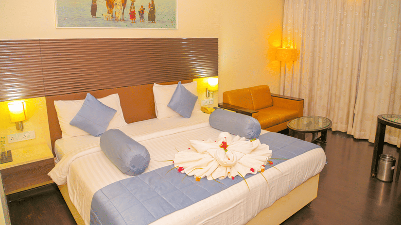 A hotel bedroom at Ambar Sarovar Portico, Gandhidham shows a large king size bed decorated with folded towels, a sofa, and a wall painting.