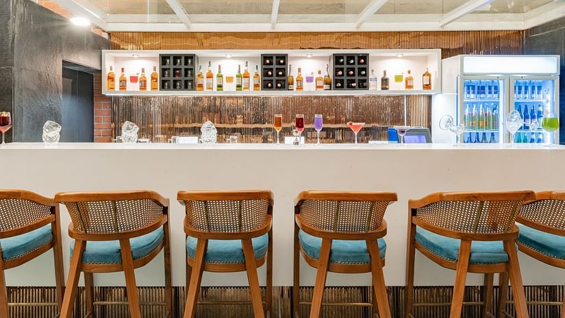 A bar with a white counter, a row of wooden stools with blue cushions, and a shelf displaying bottles of liquor - Clarks Inn, Indore