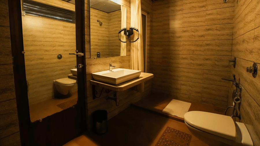 A warmly lit washroom at Adamo The Village featuring a wall mirror, countertop basin, and neatly arranged amenities.