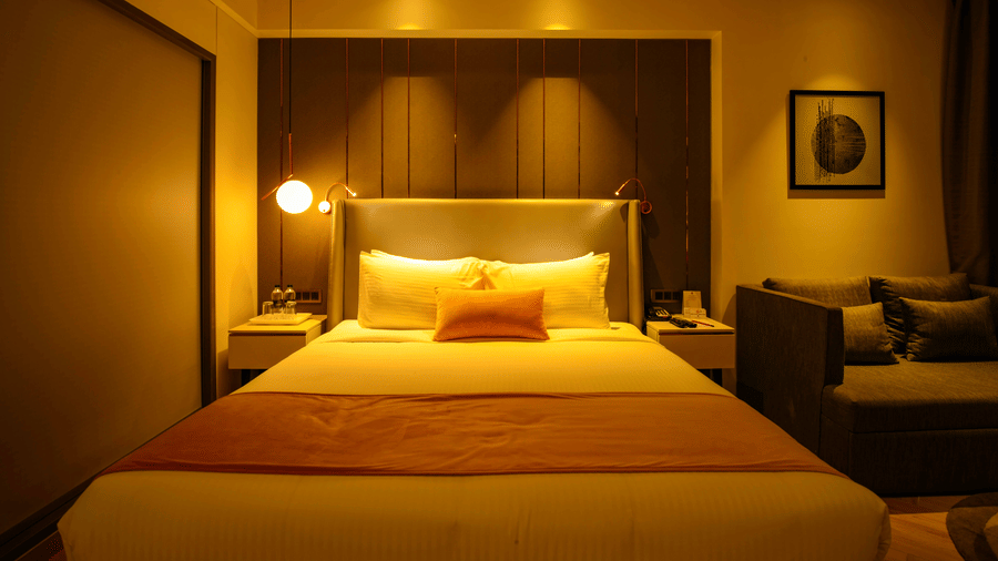 A luxurious hotel room with a plush king-sized bed, golden lighting, bedside lamps, and a cosy seating area with a sofa - Hotel Kanha Shyam, Prayagraj