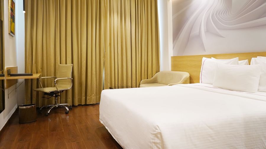 A corner view of a spacious guest room at Nemesia featuring a comfortable bed, a work desk, a seating area, and floor-to-ceiling curtains, creating a relaxing and inviting atmosphere.