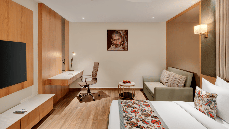 A hotel suite with a bed featuring a floral runner, a sofa seating area with a low table, light wood panelling on the walls, and a television | Voyage Eastin Suites