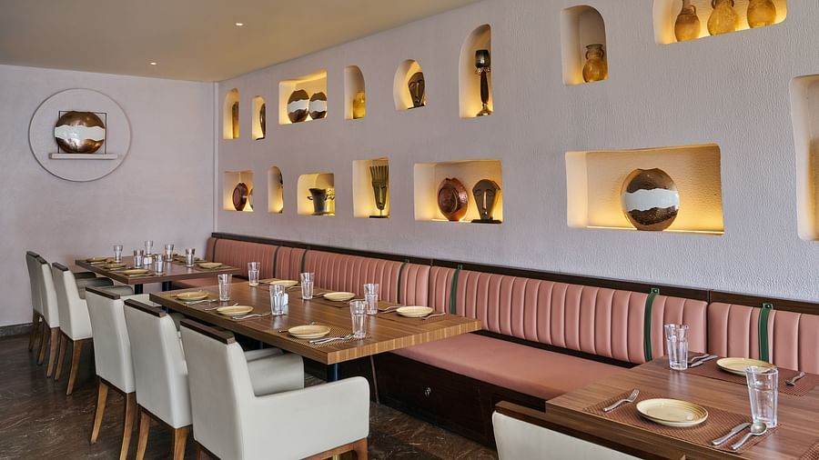 A restaurant dining area shows tables set with plates and glasses, positioned next to a long banquette with pink upholstery. The textured white wall is decorated with illuminated niches displaying various decorative objects - Voyage The Solitaire Bliss.