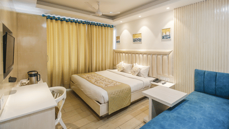A well lit hotel room featuring a queen size bed with a decorative runner, a blue sofa, a small table, a desk with a chair, and a window with yellow curtains - Diamond Hotels