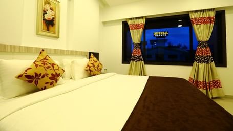 A king size bed with white and brown bedding in the suite room, featuring a painting hanging above the bed, and a window with patterned curtains at Imperial Grande.