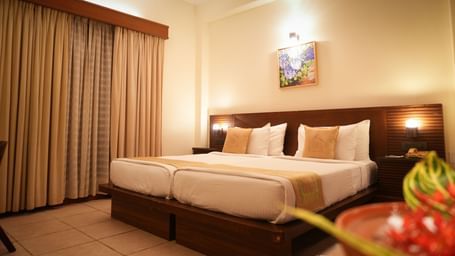 A warmly lit Superior Room at Vedic Village Spa Resort featuring a double bed with a wooden headrest, drapes over a large window, and a painting above the bed.