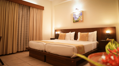 A warmly lit Superior Room at Vedic Village Spa Resort featuring a double bed with a wooden headrest, drapes over a large window, and a painting above the bed.