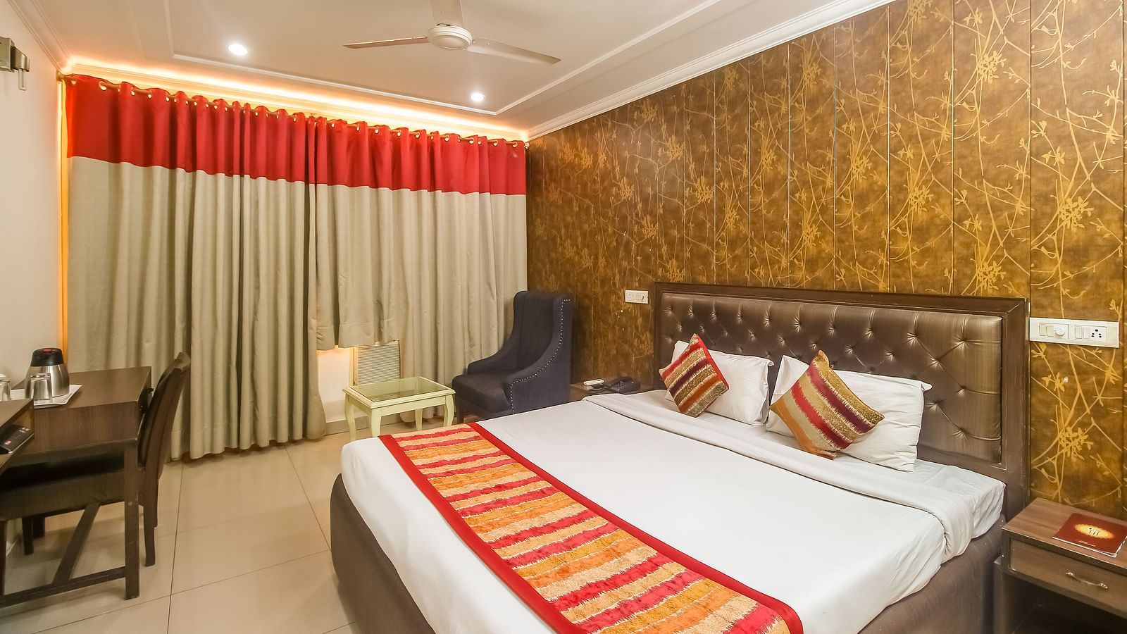 A hotel room with a textured accent wall, a plush bed with decorative pillows and a runner, a dark armchair, and a desk, illuminated by warm lighting. - Hotel Diamond Plaza, Chandigarh.