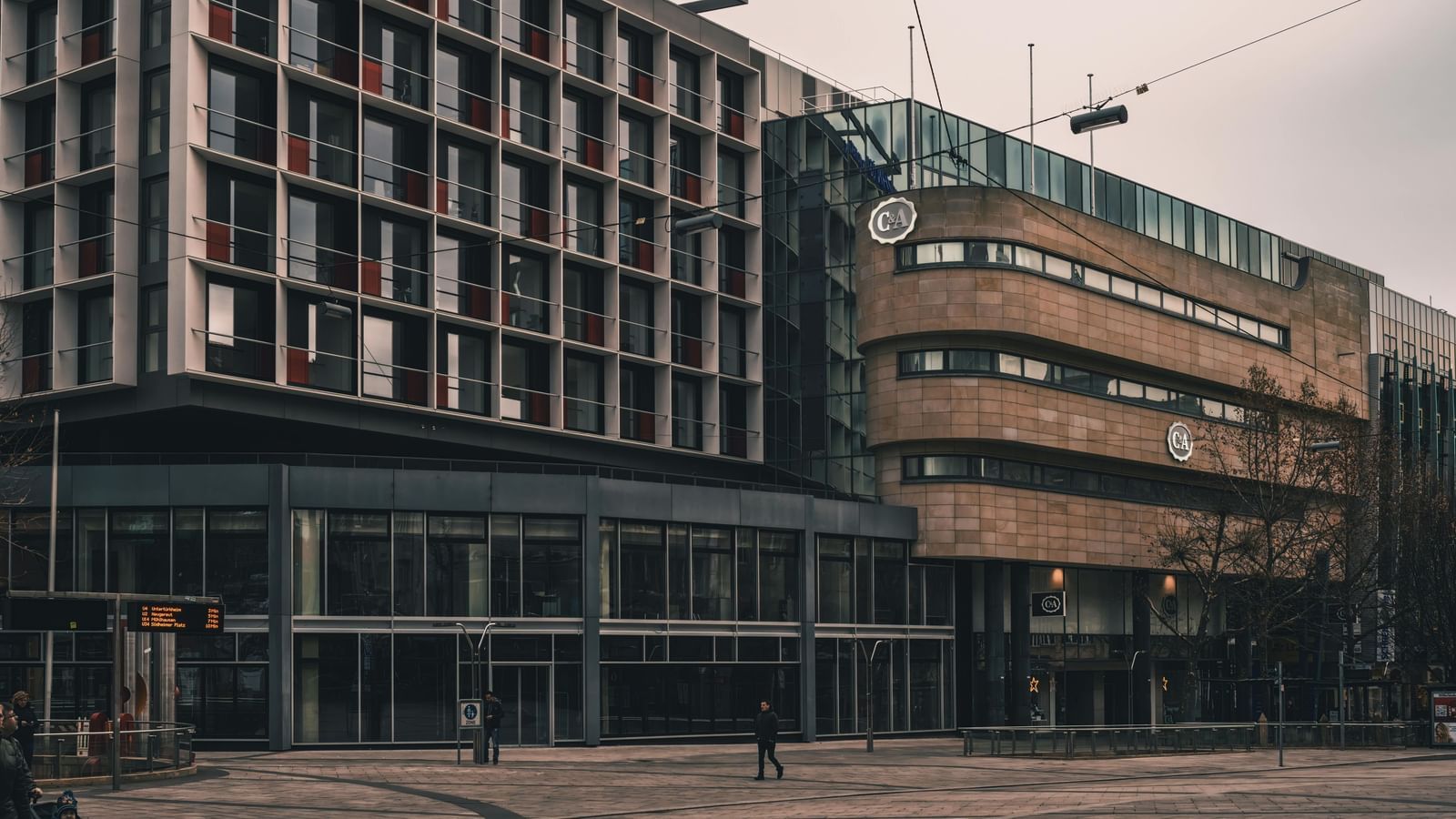 A wide-angle, low-angle shot of two modern buildings, one with a grid of windows and the other with curved, layered facades, under a grey sky.