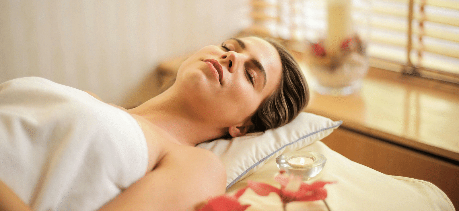 A woman with her eyes closed lying down on a spa bed alongside a small candle.