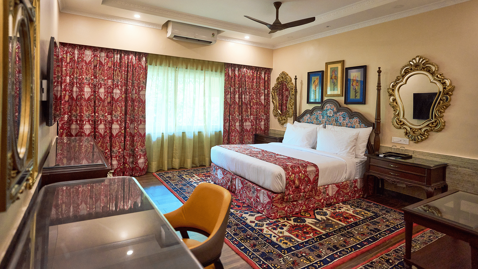 A spacious room featuring a large bed, a desk with a chair, wooden bedside tables, and windows draped with curtains at Mayfair Manor, Alibag