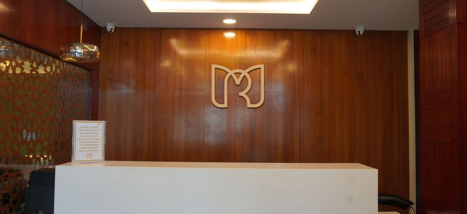 Reception area of Mertiya Residency with a minimalist white counter, warm wooden wall panels, and a stylish illuminated logo in the background.