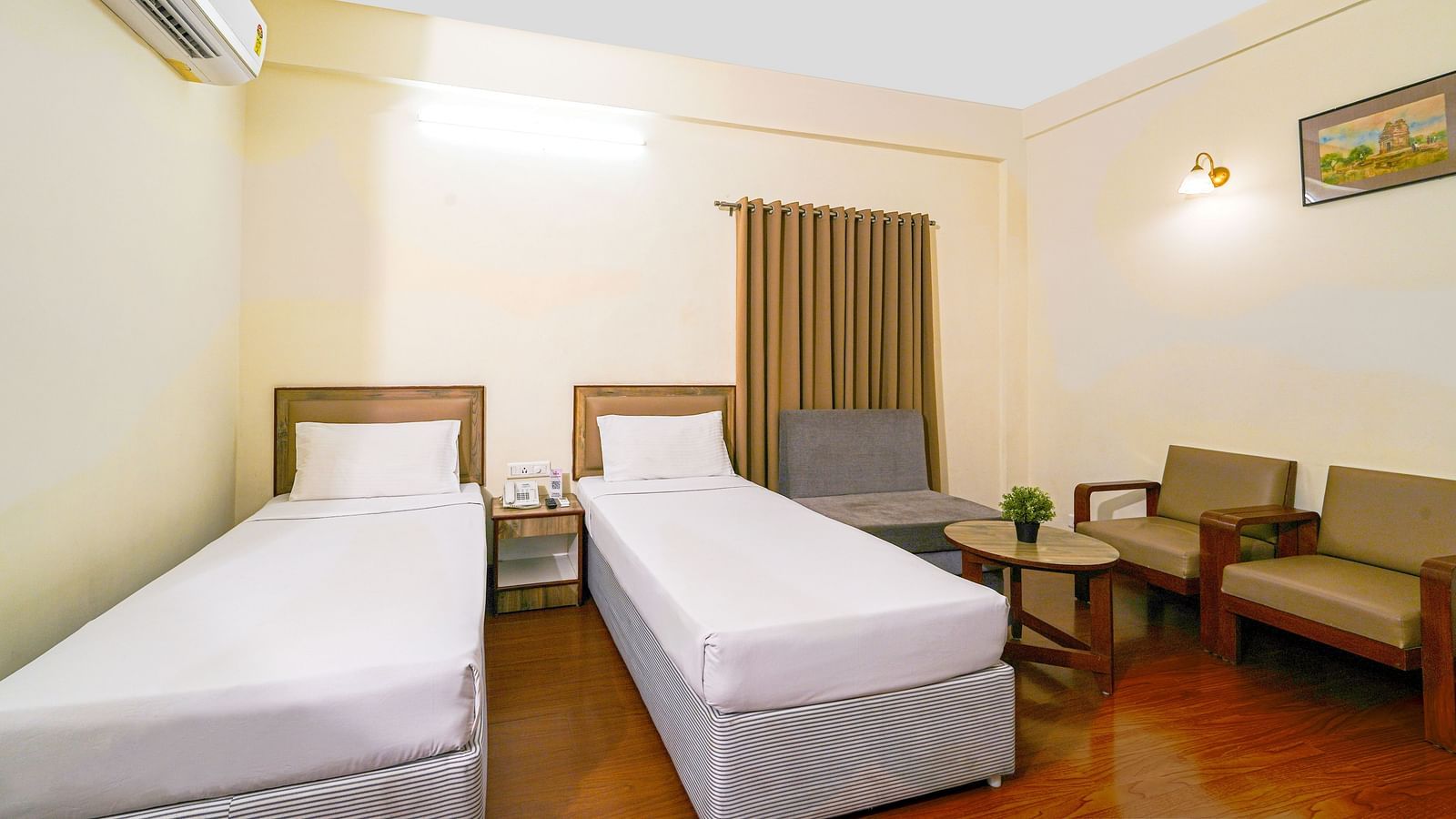 Well-lit hotel room with two single beds, wooden flooring, and a spacious seating area in Hyderabad near Ramoji Film City.
