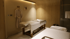 A spa treatment room featuring two massage tables covered in white linens, soft ambient lighting, wooden walls and ceiling, two bathrobes hanging on the wall, a towel on a rack, and a framed artwork above the robes - Golden Tulip, Lucknow