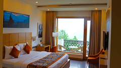 A bright hotel room with a large bed, featuring a wooden headboard and an orange and patterned decorative runner.
