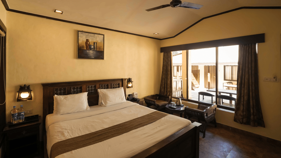 A room at Adamo The Village with a double bed, a large window, a seating area, and a side table with a lamp.