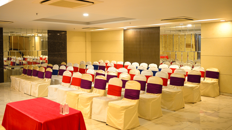 Rows of fabric covered chairs face forward inside a carpeted seminar room setup at Ambar Sarovar Portico, Gandhidham with a covered table.