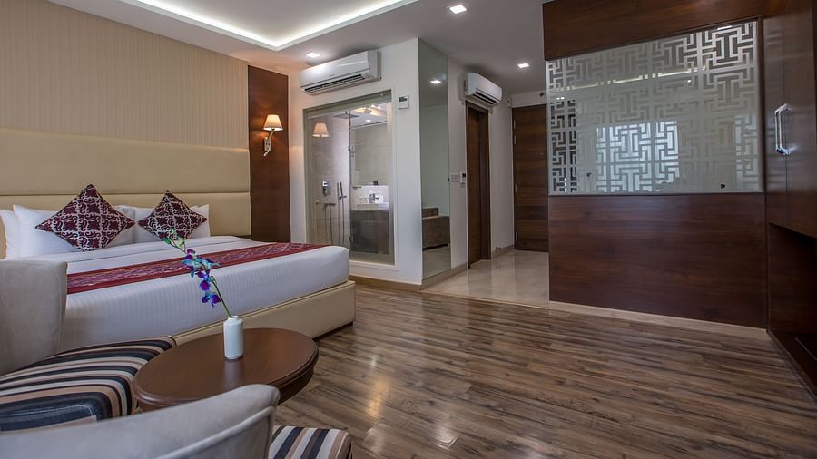 Executive Suite at Clarks Inn Suites, Near Gwalior DD Mall, featuring a king-size bed with patterned cushions, wooden flooring, glass-walled bathroom, wall-mounted lights, seating area with striped chairs, and decorative frosted glass partition.