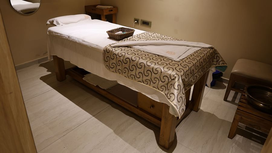 Massage room with a neatly made bed, patterned blanket, wicker basket, mirror on the wall, wooden furniture, and soft ambient lighting - Golden Tulip, Lucknow
