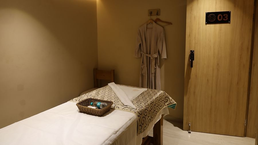 Spa treatment room with a massage bed, robe on a hook, ambient lighting, wooden door labeled “03,” and a wicker tray with a water bottle and toiletries - Golden Tulip, Lucknow