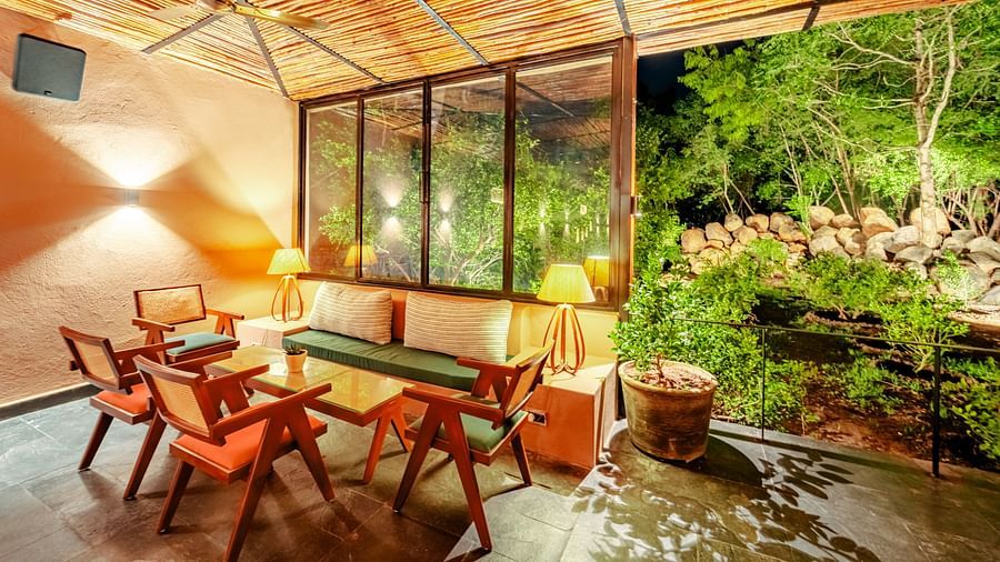An image showing wooden chairs and table placed aesthetically inside a room with bamboo roofing, where the garden view is at an enjoyable distance
