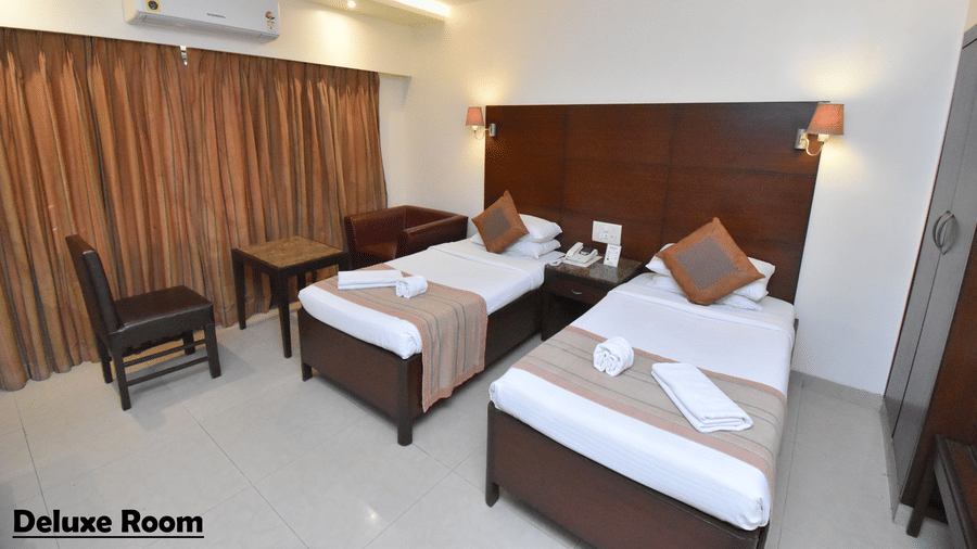 A Deluxe Room at Kings International Juhu featuring 2 twin beds, 2 chairs facing a small table, an A/C unit and a tiled floor. 