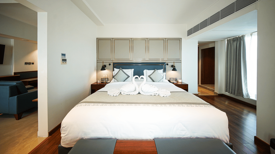An interior view of a bedroom section within the Premium Suite Room at Pride Premier Udaipur. A double bed with pillows and a folded fabric arrangement sits in the centre of the room, positioned between bedside units.