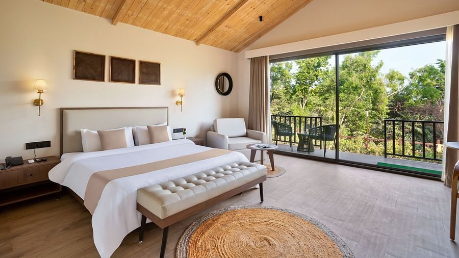 An image of a premium cottage featuring a king size bed, a bedside table and lamp on either side, a sofa chair, and a spacious balcony attached to the room at Stone Woods Premier Resort, Gokarna